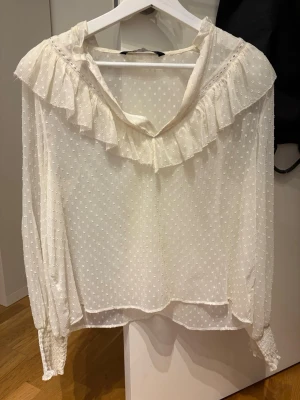 Vit blus med volangkrage - Romantisk vit blus med volangkrage och diskret prickigt mönster. Blusen har långa ärmar med smock vid ärmsluten och är tillverkad i ett tunt, lätt och transparent tyg. Perfekt för en trendig och feminin look.