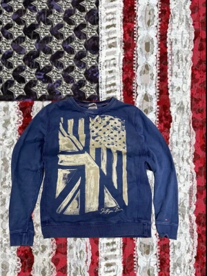 Blå sweatshirt från Hilfiger Denim - Blå sweatshirt från Hilfiger Denim med stort tryck av amerikanska och brittiska flaggan i silver på framsidan. Rund halsringning och långa ärmar. Perfekt för dig som gillar streetwear och vill sticka ut med en unik design. Passar S/M.