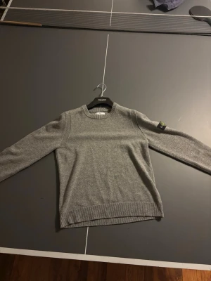 Grå stickad tröja från Stone Island - Snygg grå stickad tröja från Stone Island med klassisk rund hals och ribbade muddar vid ärmslut och nederkant. Köpte den på Nk för cirka ett år sedan.Tröjan har den ikoniska Stone Island-loggan på vänster ärm och är perfekt för dig som gillar stilrena plagg. Redo att gå ner i pris för snabb affär, 