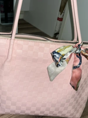 Rutig rosa handväska med scarfdetalj - Snygg rosa handväska i rutigt mönster med dragkedja upptill och två långa handtag. Väskan har en färgglad scarf knuten på ena handtaget som extra detalj. Perfekt för dig som vill ha en trendig och rymlig väska med unik touch.