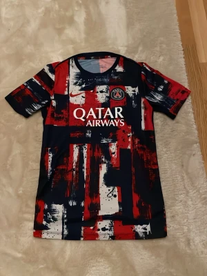 PSG Nike Dri-Fit träningströja XS - Snygg PSG träningströja från Nike med Dri-Fit material. Tröjan har ett unikt abstrakt mönster i rött, marinblått och vitt samt PSG-logga och Qatar Airways-tryck på bröstet. Perfekt för fotbollsträning eller sportiga dagar. Köpt i Nike store i London. Original pris 599kr 