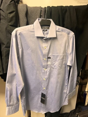 Blå skjorta från Ralph Lauren - Helt ny Klassisk ljusblå skjorta från Ralph Lauren i slim fit-modell som aldrig har kommit till användning då det var en present i  för stor storlek. Skjortan har långa ärmar, bröstficka och vita knappar framtill. Tillverkad i bomull och har en stilren, tidlös look som passar perfekt till många olika outfits.