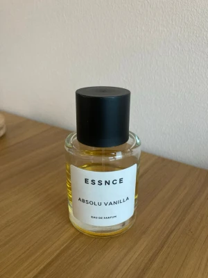 ESSNCE Absolu Vanilla Eau de Parfum - ESSNCE Absolu Vanilla Eau de Parfum i en rund glasflaska med svart matt kork. Parfymen har en varm gul vätska och en stilren vit etikett med svart text. Doften har tydliga vaniljtoner och flaskan ger ett minimalistiskt och modernt intryck.