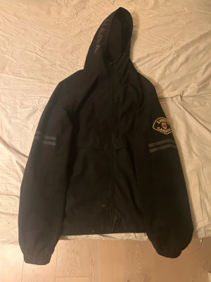 Svart vindjacka från Zanetti Canada - Svart vindjacka från Zanetti Canada med huva och dragkedja framtill. Jackan har ett stort märke på ärmen och en röd lönnlöv-detalj på insidan. Två diskreta ränder på ärmarna och elastiska muddar. Perfekt för dig som vill ha en stilren och funktionell jacka. Har några små fläckar men inget som man lägger märket till 