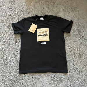 Svart Burberry t-shirt med beige patch - Svart t-shirt från Burberry med beige fyrkantig patch och svart tryck på bröstet. Klassisk rund hals och korta ärmar. Stort Burberry-tryck och globmotiv framtill. Tillverkad i mjuk bomull för en skön känsla.