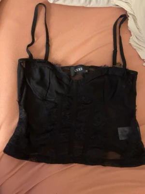 Svart spetsig korsettopp från Cider - Snygg svart korsettopp från Cider med spetsdetaljer och justerbara axelband. Toppen har en figurnära passform och genomskinliga partier i spets, perfekt för att lyfta din outfit.