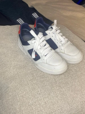 Vita och marinblå sneakers från lyle and Scott - Snygga sneakers från Lyle and Scott i vitt och marinblått med röda detaljer. Skorna har klassisk snörning, rund tå och platt sula. Tillverkade i skinn och syntetmaterial med logga på sidan och dragflik bak.