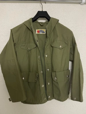 Grön vindjacka/overshirt från Fjällräven - Snygg grön vindjacka från Fjällräven med huva och flera praktiska fickor framtill. Jackan har dragkedja och tryckknappar, samt en patch med logga på ärmen. Perfekt för dig som gillar outdoor och stilren design.
