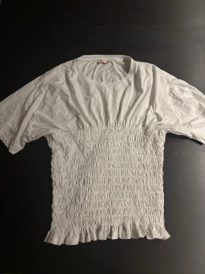 Vit smockad topp från Nelly - Supersöt vit topp från Nelly med smockad midja och volangkant nedtill. Toppen har korta ärmar och rund halsringning, vilket ger en avslappnad men ändå feminin look. Perfekt att styla med jeans eller kjol för en trendig outfit.