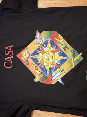 Casa blanca t-shirt - Svart t-shirt från Casa Blanca med stort färgglatt tryck av sol, fåglar och flygplan på ryggen. Framsidan har samma motiv i mindre format och texten 'CASA BLANCA' i rött och grönt. Rund halsringning och rak passform.