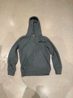 Grå zip hoodie från Superdry - Mörkgrå hoodie med dragkedja från Superdry. Klassisk modell med huva och dragsnören, två fickor framtill och broderad Superdry-logga på bröstet. Tillverkad i mjukt sweatshirtmaterial som är perfekt för chill dagar.