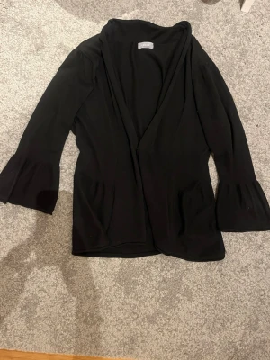 Svart kofta med trumpetärmar från ZARA - Stilren svart kofta från ZARA med vida trumpetärmar och öppen front. Koftan har två fickor framtill och är gjord i ett mjukt, tunt material som faller snyggt. Perfekt att slänga över en topp för en enkel men trendig look.