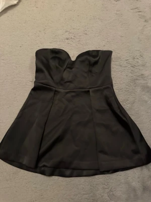 Svart axelbandslös topp - Säljer en svart topp i satinliknande material med hjärtformad urringning. Klänningen har en lätt utsvängd kjol och kan bäras både med och utan tunna axelband. Perfekt för dig som vill ha en enkel men snygg look.