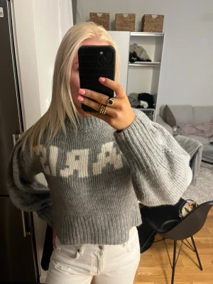 Grå stickad tröja med PARIS-tryck - Mysig grå stickad tröja med vit text 'PARIS' framtill. Tröjan har en croppad passform, ribbade muddar och lång ärm. Perfekt för dig som gillar en avslappnad men trendig look.