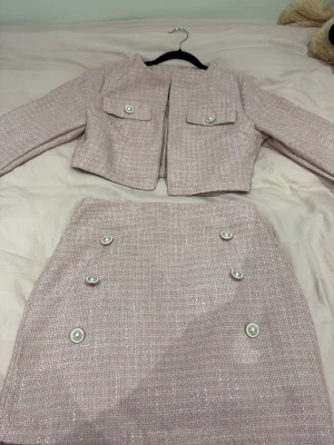 Rosa tweed set med kjol och jacka - Säljer ett stilrent rosa tweed set med matchande kjol och kort jacka. Setet har dekorativa pärlknappar och fickdetaljer på jackan. Materialet är strukturerat och skimrar lätt. Perfekt för dig som gillar en chic och trendig look. Färgen ser tråkig ut på bilden, men den är en härlig rosa färg