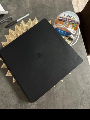 PlayStation 4 Slim svart konsol och vit - Säljer en svart PlayStation 4 Slim från Sony. Konsolen har en stilren, fyrkantig design i matt plast och kommer med spelet WRC 6 för PS4. Perfekt för gaming och underhållning hemma. 