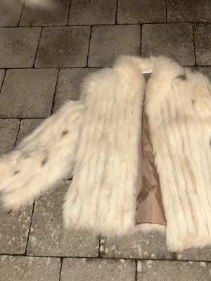 Vit pälsjacka i rävpäls från Finland - Säljer en fluffig och vit pälsjacka i äkta rävpäls med beige foder i polyester. Jackan har en klassisk rak passform och lång ärm. Räven är från Finland. Vet tyvärr inte årgången på den då jag fick den från en äldre släkting 