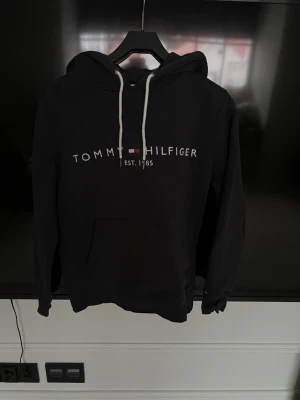 Svart hoodie Tommy Hilfiger - Svart hoodie från Tommy Hilfiger med vit dragsko i huvan och klassisk logga samt texten 'EST. 1985' på bröstet. Stor känguruficka framtill och mjukt material som känns skönt mot huden. Perfekt för en avslappnad och stilren look.