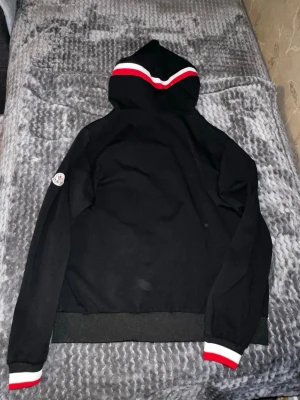 Svart Moncler hoodie med dragkedja - Snygg svart hoodie från Moncler med dragkedja framtill och huva. Detaljer i rött och vitt på huva och ärmslut samt Moncler-logga på ärmen. Tillverkad i mjukt material som känns skönt mot huden. Perfekt för en avslappnad och trendig look. OBS DEN KÄNNS SOM STORLEK M OCH PRIS ÄR EJ HUGGET I STEN