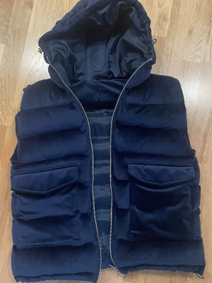 Vendiera väst (limited editon) - Snygg mörkblå dunväst från Vendiera med stor vadderad huva och två rejäla framfickor. Västen har en quiltad design och dragkedja framtill. Materialet är mjukt och ger en lyxig känsla. Perfekt för lager på lager och streetwear-stil. Storlek L men passar även M