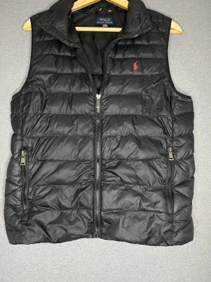 Svart dunväst från Polo Ralph Lauren - Snygg svart dunväst från Polo Ralph Lauren med klassisk röd logga på bröstet. Västen har två dragkedjeförsedda fickor och quiltad design. Perfekt för lager-på-lager och håller dig varm med lätt dunfyllning. Stängs med dragkedja framtill.