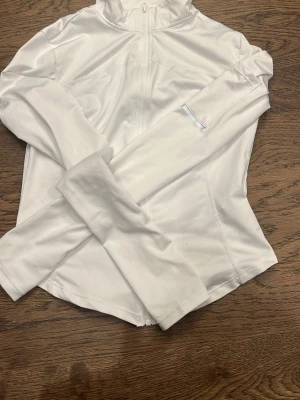 Vit träningströja med ficka - Snygg vit ziptröja i stretchigt material med dragkedja framtill. Tröjan har en praktisk ficka på ärmen med en reflekterande detalj, perfekt för träning eller chill. Figurnära passform och långa ärmar.