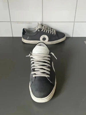 Svarta sneakers från O2 Oliver Cabell - Snygga svarta sneakers från Oliver Cabell med vit sula och vita snören. Skorna har en rund tå och ett stort vitt O på sidan som detalj. Tillverkade i skinn med klassisk låg profil och platt sula. Perfekta för en clean och stilren look.