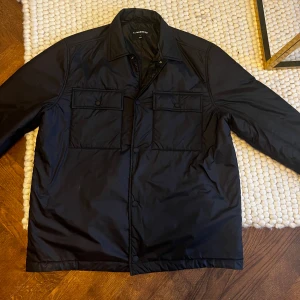 Svart dunovershirt från J.Lindeberg - Stilren svart dunjacka från J.Lindeberg med klassisk krage, två stora bröstfickor med lock och knappar samt diskret knappstängning framtill. Jackan är tillverkad i glansig polyester och har en rak, normal passform med långa ärmar.