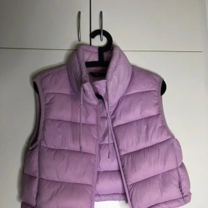Ljuslila dunväst från Zara - Säljer en ljuslila dunväst från Zara med hög krage och dragsko. Västen är quiltad och har en croppad passform som ger en trendig look. Perfekt att slänga över en hoodie eller långärmad tröja för extra värme.