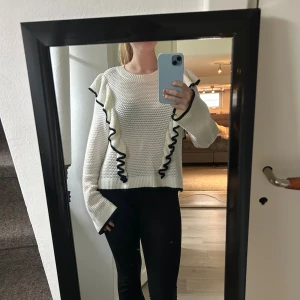 Vit stickad tröja med svarta volanger - Säljer en vit stickad tröja med svarta volangdetaljer längs framsidan och ärmsluten. Tröjan har en rund halsringning och långa ärmar med lite vidare muddar. Perfekt för dig som vill ha något unikt och snyggt i garderoben! 🩷Strl: M men passar definitivt S🩷