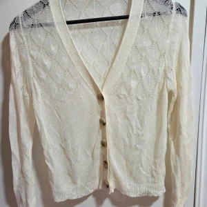 Vit stickad kofta med knappar - Söt vit stickad kofta med lång ärm och v-ringning. Koftan har ett diskret rutmönster i spets och stängs med fem stora knappar framtill. Perfekt att slänga över en topp för en chill look. Materialet känns lätt och luftigt.