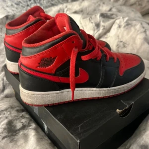 Nike Air Jordan 1 Mid Svart/Röd 38.5 - Säljer ett par Nike Air Jordan 1 Mid i svart och rött skinn med klassisk röd swoosh och snörning. Skorna har vit sula och ikonisk Jordan-logga på sidan. Perfekta för dig som vill sticka ut med en tidlös streetstyle💞
