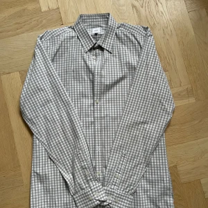 Rutig skjorta från Bläck i grått - Snygg rutig skjorta från Bläck i grått och vitt. Skjortan har klassisk krage, långa ärmar och knäppning framtill. Perfekt för dig som gillar stilrena och enkla plagg med lite edge. Tillverkad i mjuk bomull som känns skön mot huden.