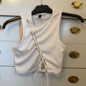 Vit ribbad linnetopp med snörning - Säljer en vit ribbad linnetopp från H&M med cool snörningsdetalj framtill. Toppen är ärmlös och croppad, perfekt till jeans eller kjol. Materialet är mjukt och stretchigt för en bekväm passform.