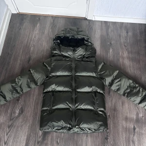 Mörkgrön pufferjacka från Everest - Säljer en mörkgrön pufferjacka från Everest med huva och två dragkedjeförsedda fickor framtill. Jackan har en glansig yta och är vadderad för extra värme. Perfekt för kalla dagar och har en snygg badge på ärmen. Den är endast använd 3 gånger och säljer pga att vill köpa en annan. Den är i storlek xs men passar mig Som har S.