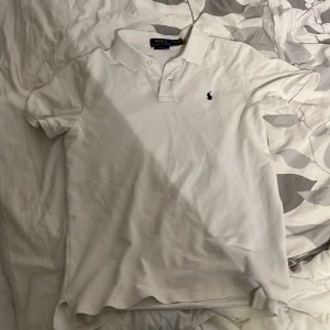 Vit pikétröja Polo Ralph Lauren - Vit pikétröja från Polo Ralph Lauren i slim fit-modell. Klassisk krage, korta ärmar och broderad marinblå logga på bröstet. Tillverkad i bomull för en skön och luftig känsla. Perfekt för en clean och stilren look. Den har en liten fläck som inte går back på ryggen, men den märks knappt av!