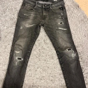 Replay Anbass svarta slitna jeans - Säljer ett par Replay Anbass jeans i svart tvätt med slitningar och hål för en riktigt edgy look. Jeansen har klassisk femficksdesign, smal passform och snygga detaljer på bakfickan. Perfekta för dig som gillar streetstyle och vill sticka ut.