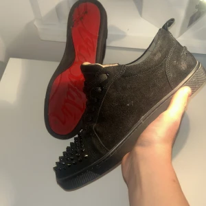 Christian Louboutin svarta sneakers med nitar - Svarta sneakers från Christian Louboutin i mocka med coola svarta nitar på tån och klassisk röd sula med logga. Skorna har snörning och rund tå, samt platt sula. Perfekt för dig som gillar edgy designerstil och vill sticka ut. Kan sänka pris vid snabb affär 