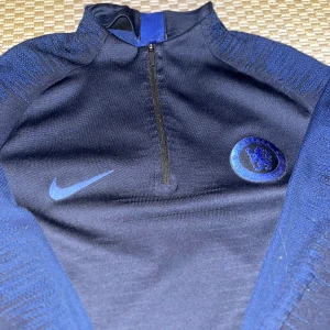 Nike Vaporknit - Chelsea - Chelsea FC långärmad träningströja från Nike med halv dragkedja. Mörkblå och blå färg, tillverkad i tekniskt VaporKnit-material för bästa andningsförmåga. Klubbmärke och Nike-logga på bröstet. Perfekt för fotbollsträning eller gym.
