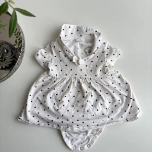 babyklänning  - Supersöt vit babyklänning från H&M i ekologisk bomull med svarta prickar. Klänningen har korta ärmar, en liten krage med knyt och inbyggd body med tryckknappar. Perfekt för små äventyr och lek!