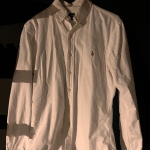 Vit skjorta från Ralph Lauren slim fit - Klassisk vit skjorta från Ralph Lauren i slim fit-modell. Skjortan har knappar hela vägen, button down-krage och den ikoniska broderade loggan på bröstet. Tillverkad i mjuk bomull och har långa ärmar. Perfekt för en clean och stilren look.