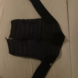 Svart dunväst från Moncler - Svart Cardigan från Moncler med quiltad framsida och två dragkedjeförsedda fickor. Cardigan har Moncler-logga på vänster ärm och ribbade partier på sidorna. Stängs med dragkedja framtill och har en stilren, sportig look som passar perfekt till höst och vinter.
