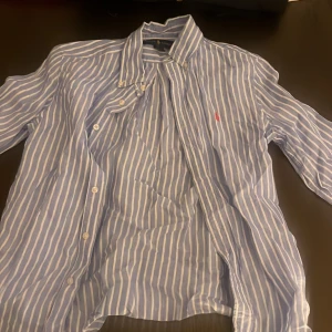 Blåvit randig skjorta från Ralph Lauren - Snygg blå och vit randig skjorta från Ralph Lauren med klassisk krage och knäppning framtill. Skjortan har långa ärmar och den ikoniska röda loggan broderad på bröstet. Perfekt för en stilren och avslappnad look.