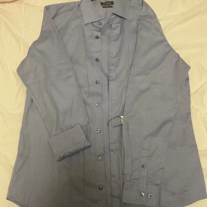 Blå skjorta Dressmann XL - Helt ny Snygg blå och vit randig skjorta från Dressmann i slim fit-modell. Tillverkad av exklusiv egyptisk bomull som känns mjuk och lyxig. Skjortan har långa ärmar, klassisk krage och knäppning framtill. Perfekt för dig som gillar stilrena och fräscha plagg.