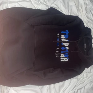 Svart Trapstar hoodie med blå/vit text - Svart hoodie från Trapstar London med fet blå och vit text på bröstet. Klassisk känguruficka, huva med snörning och mjukt material på insidan. Perfekt för dig som gillar streetwear och vill sticka ut med en snygg logga.