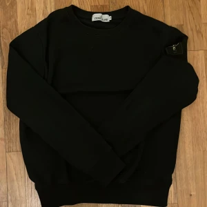 Svart sweatshirt från Stone Island - Svart sweatshirt från Stone Island med klassisk rund hals och ikonisk logotyp-patch på vänster ärm. Tröjan har en clean look och är tillverkad i mjuk bomull, perfekt för dig som gillar streetwear och stilrena plagg.