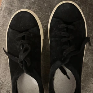 Svarta mockasneakers från & Other Stories - Svarta sneakers i mocka från & Other Stories med vit, platt sula och klassisk snörning. Skorna har rund tå och en stilren look som passar till många outfits. Tillverkade i Portugal och har en mjuk känsla tack vare mockamaterialet.