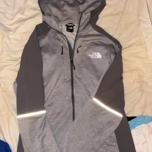 Grå tracksuit från The North Face - Snygga grå tracksuit från The North Face med reflexdetaljer vid knäna och logga på benet. Byxorna har dragkedjefickor och är tillverkade i mjukt material som passar perfekt för chill eller träning. Raka ben och bekväm passform. En fläck från blå målarfärg på byxorna som är svår att se men den finns där därav det billiga priset