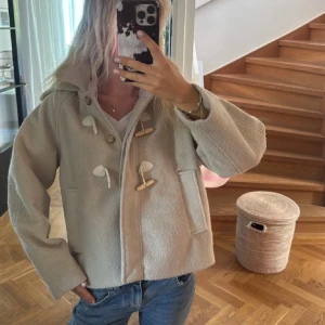 Beige duffeljacka från Zara med huva - Beige duffeljacka från Zara med huva och snygga träknappar med snörning. Jackan har två fickor fram, klassisk krage och är tillverkad i mjukt ullblandat material. Perfekt för kyliga dagar och ger en clean och stilren look.