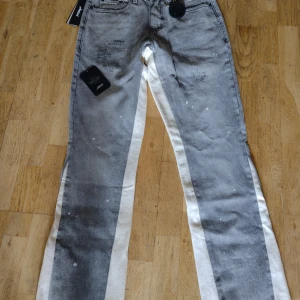 Grå jeans med vita sidor från Amicci - Snygga, helt nya grå jeans från Amicci med breda vita paneler längs sidorna och coola färgstänk-detaljer. Klassisk femficksmodell, lätt utställda och svart knapp framtill. Jeansen har en streetig vibe och är tillverkade i denim. Köpte fel storlek, så de är för stora. Betalt 1434 kr med tullavgift.